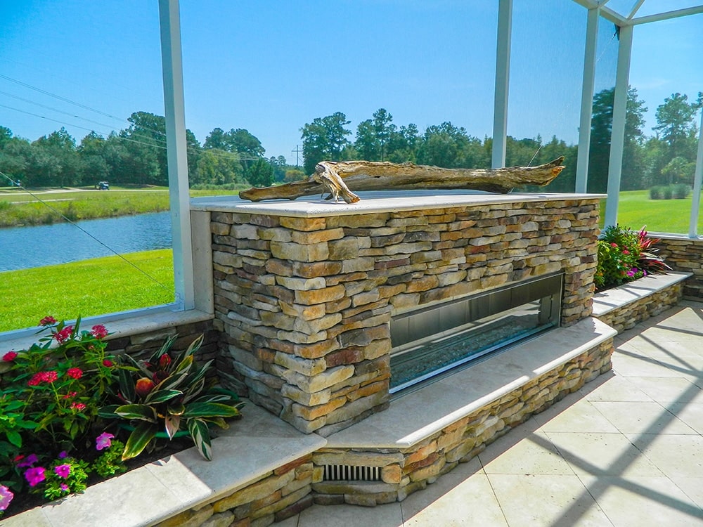 Screened outdoor living area featuring a stacked stone fireplace with a linear gas fire feature overlooking a peaceful pond and landscaped backyard.